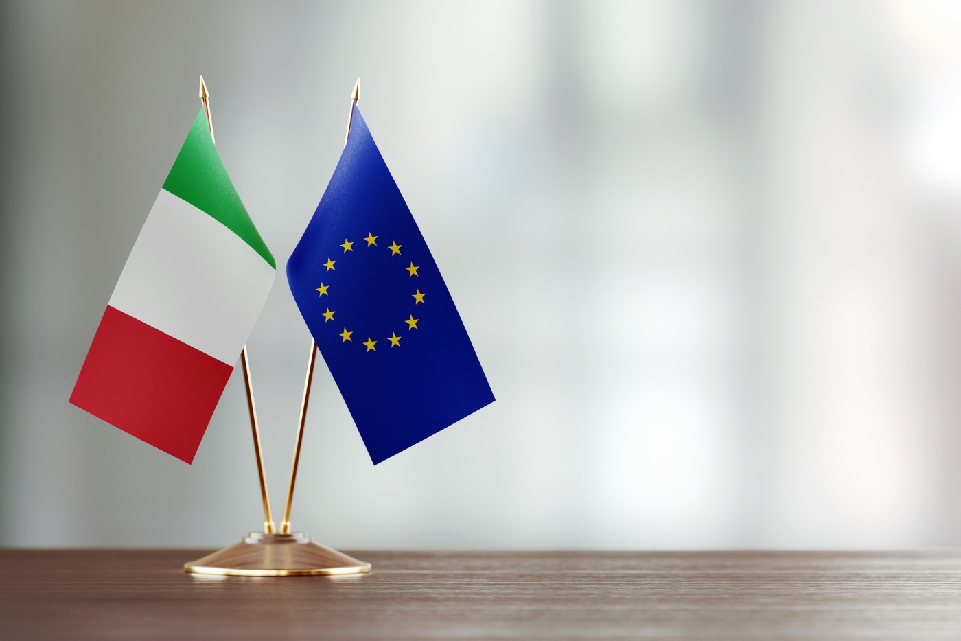 italian-and-european-union-flag-pair-on-a-desk-over-defocused-picture-id925871346